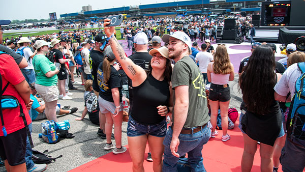 A large crowd gathers on the track at a speedway during a fan event. In the foreground, a smiling couple stands on a red carpet taking a selfie, with grandstands and event displays visible in the background.