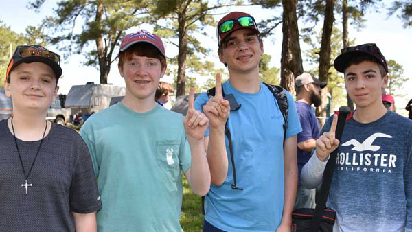 Four teenage boys pose together outdoors at a campsite, smiling and holding up one finger. They wear casual summer clothes and baseball caps, with tents and trees in the background.